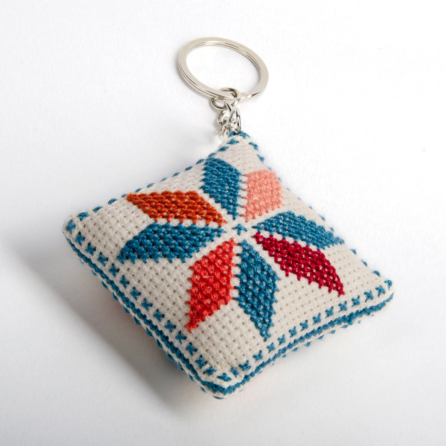 angle view of a handmade Palestinian Tatreez keychain (6x6 cm) featuring traditional embroidery in blue, orange, and sky colors on a small square pillow.