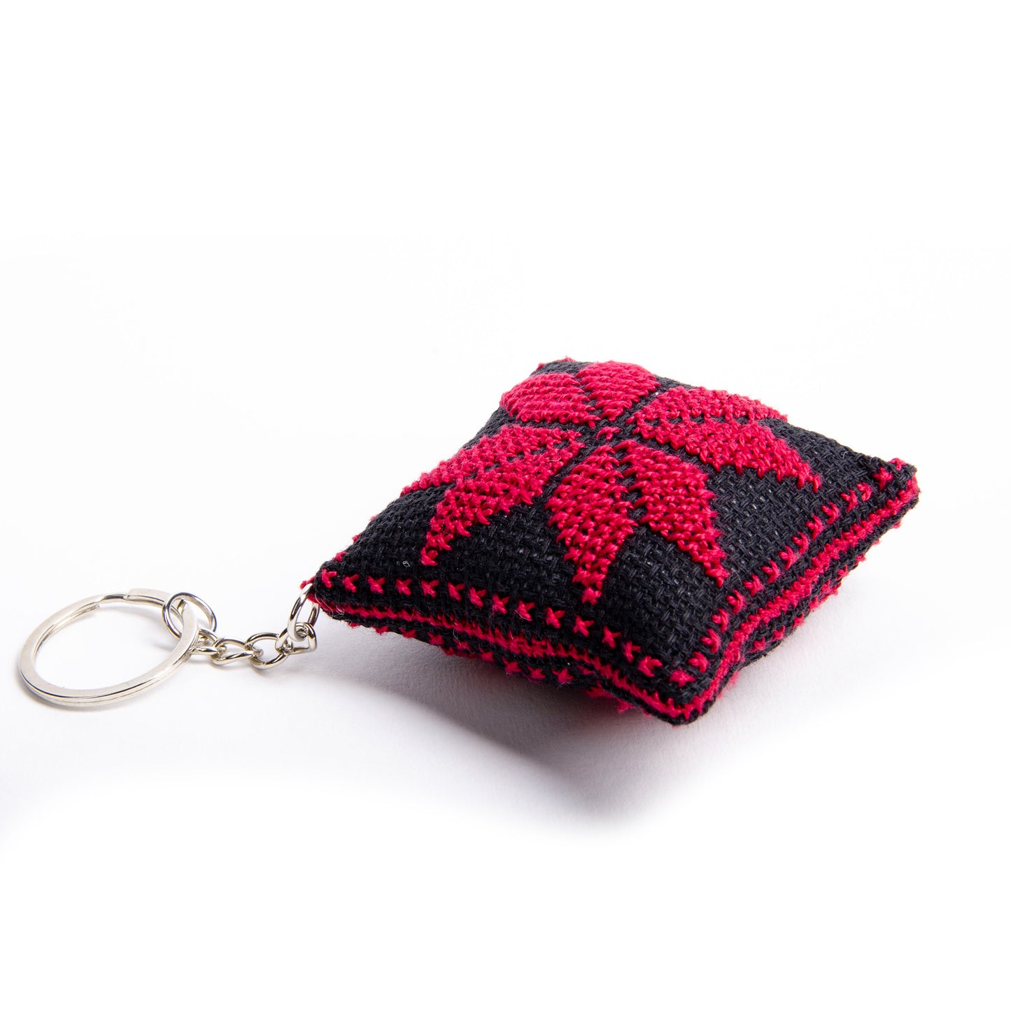 angle view of a handmade Palestinian Tatreez keychain (6x6 cm) featuring traditional embroidery in balck and red
colors on a small square pillow.