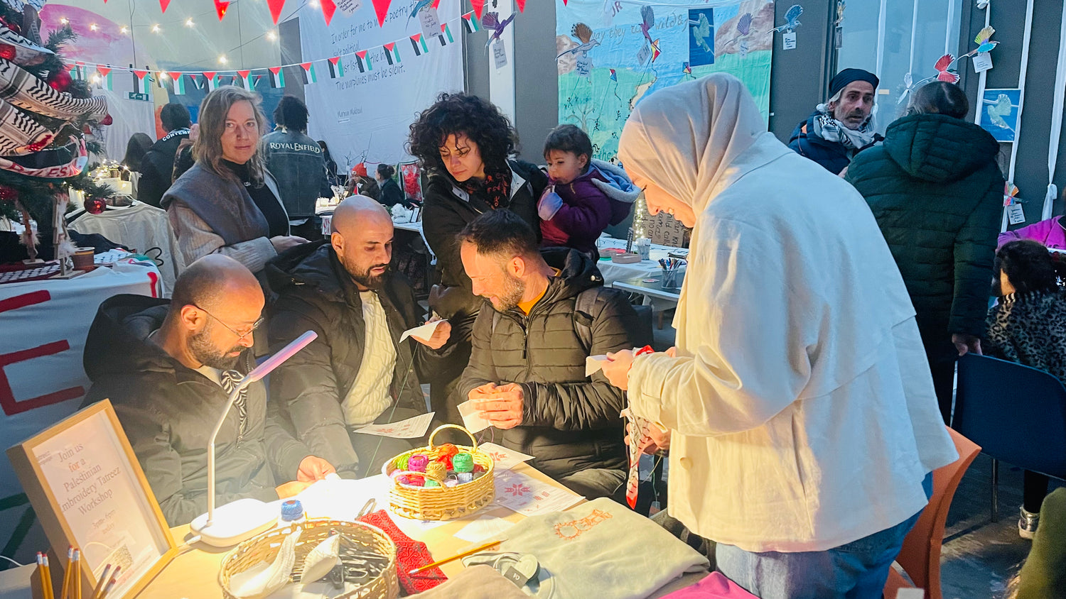 Festive Christmas setting with a bustling crowd; three men focused on creating Palestinian Tatreez as others observe, guided by a smiling instructor.