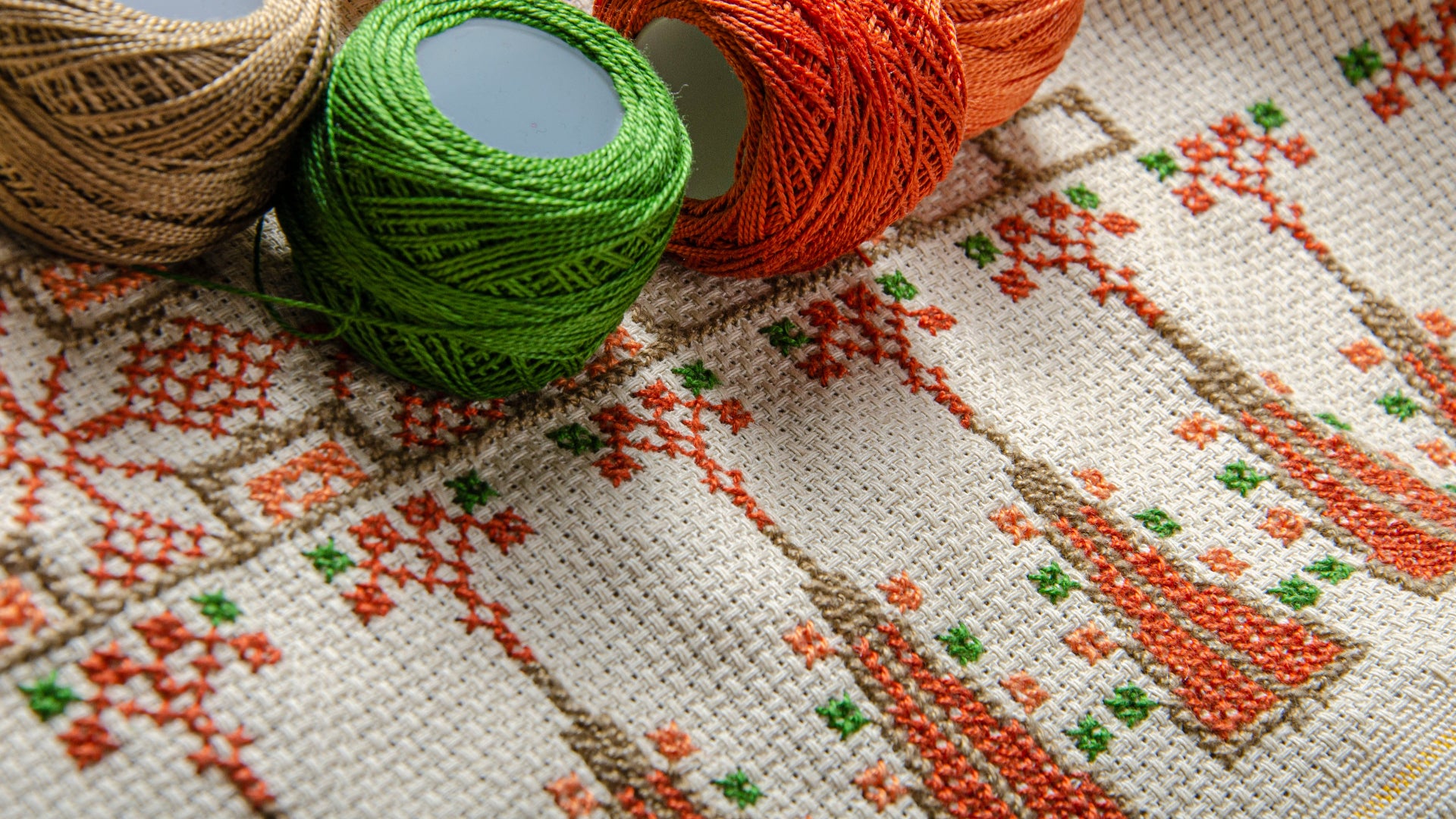 Close-up view of Tatreez fabric with orange and green patterns, accompanied by four balls of colorful thread