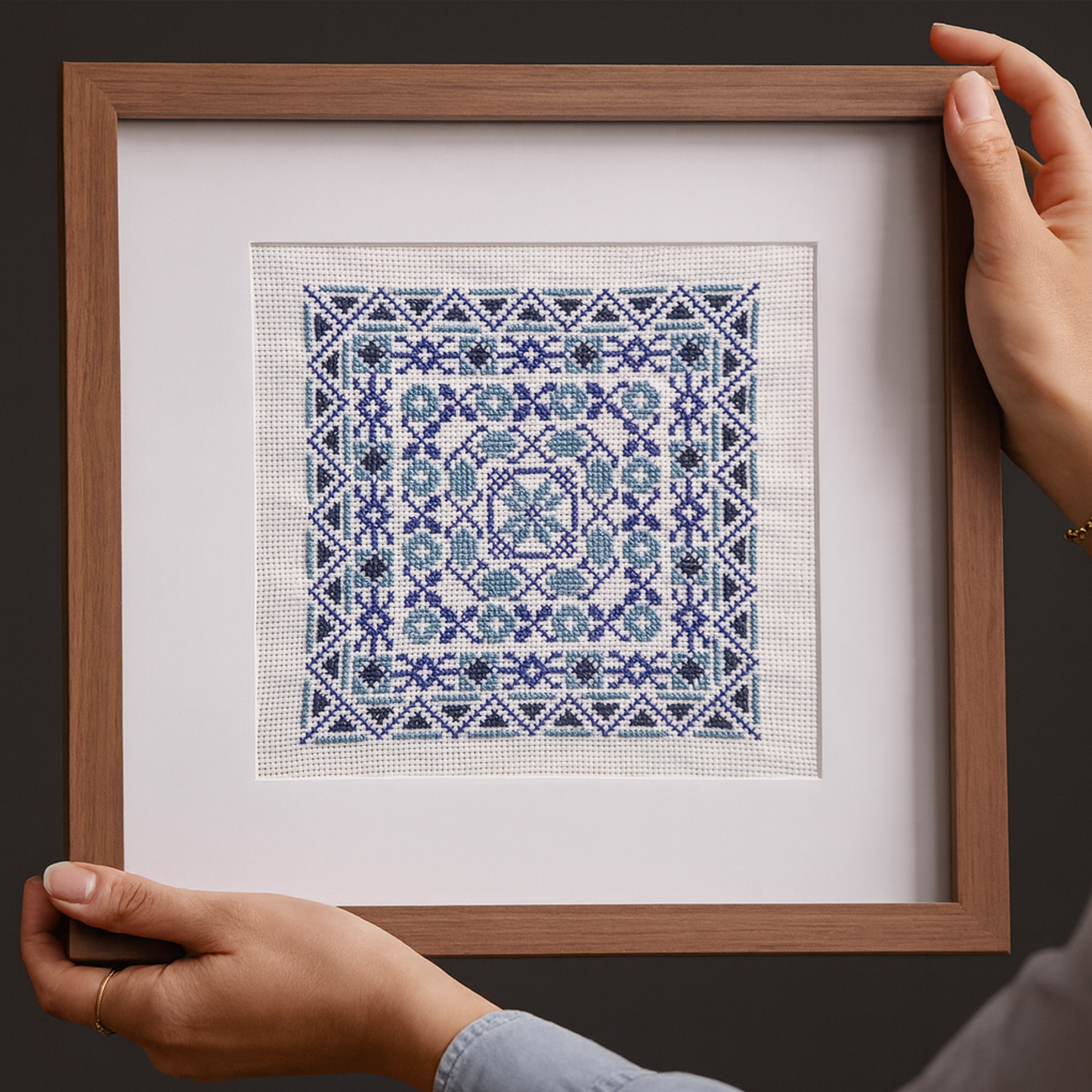 Hands holding a framed piece of Tatreez embroidery featuring an intricate geometric pattern in shades of blue and white, displayed against a dark background.