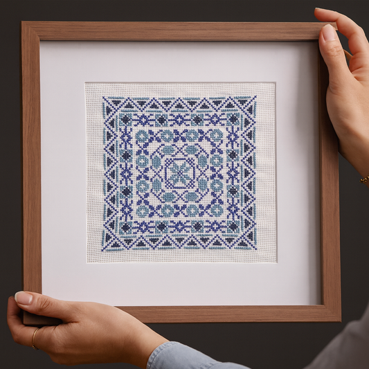 Hands holding a framed piece of Tatreez embroidery featuring an intricate geometric pattern in shades of blue and white, displayed against a dark background.
