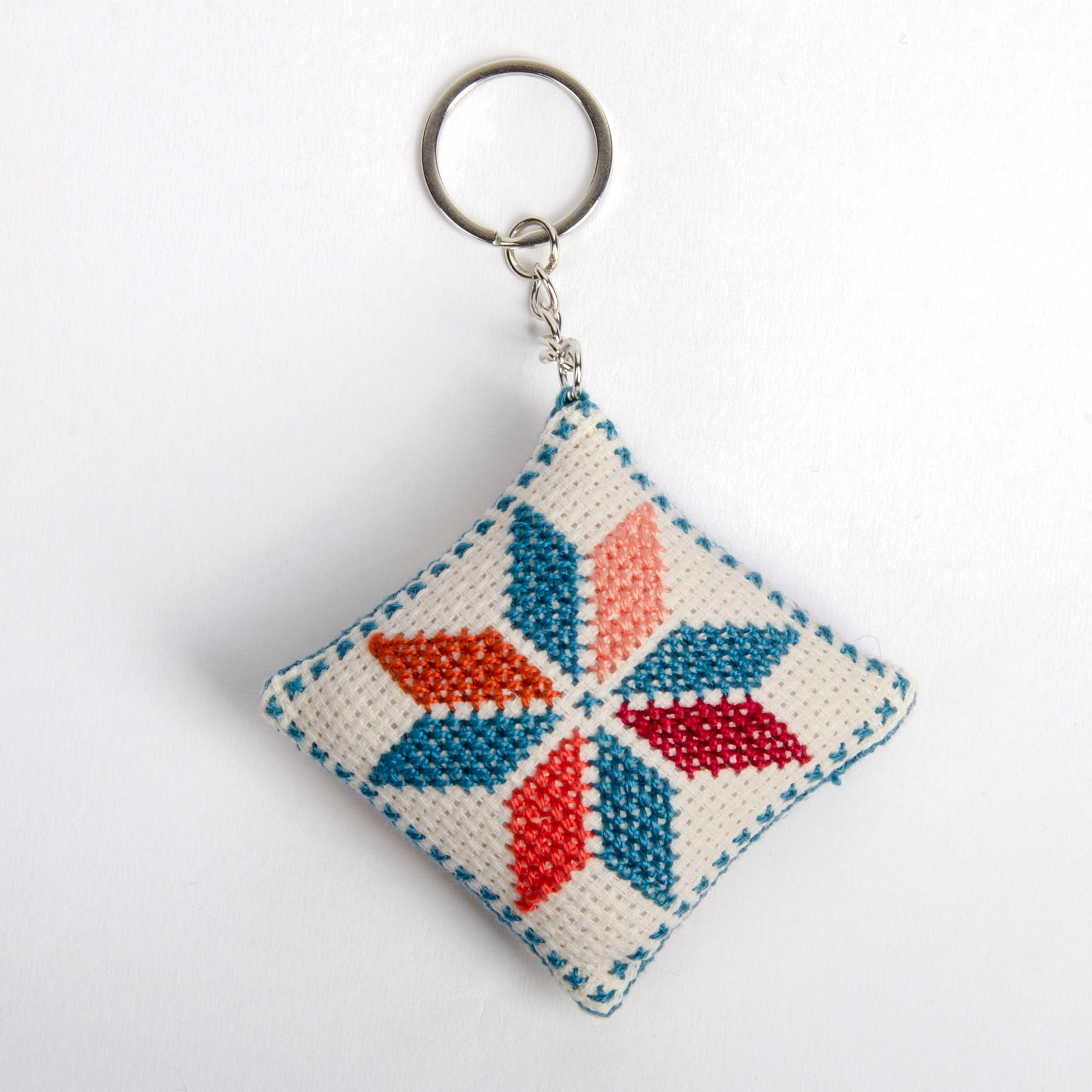 Front view of a handmade Palestinian Tatreez keychain (6x6 cm) featuring traditional embroidery in blue, orange, and sky colors on a small square pillow.