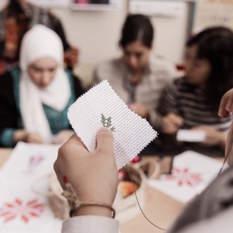 Close-up of a girl's hands stitching Palestinian Tatreez, with a blurred background of a Tatreez workshop.