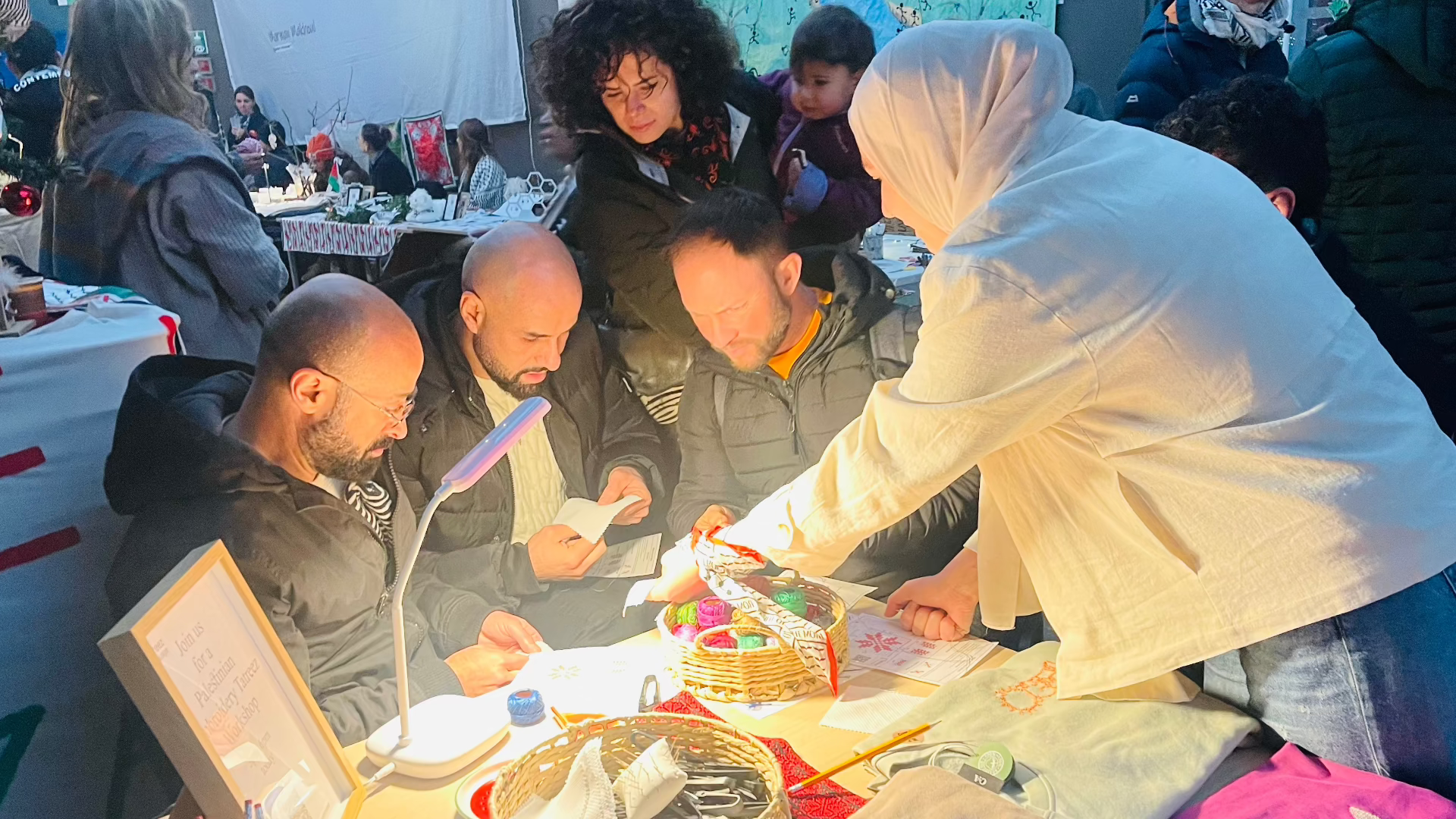 Christmas event with a lively atmosphere; three men engrossed in Palestinian Tatreez, while attendees watch and the instructor shares her expertise with joy.