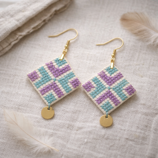 Handmade Palestinian tatreez earrings featuring the Reesh (feather) geometric motif in turquoise and purple embroidery on cream fabric, diamond-shaped with gold fishhook hooks and a small gold disc charm, photographed on natural linen with soft feathers.