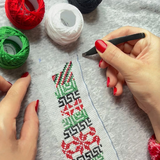 Hands stitching a traditional Palestinian tatreez pattern on fabric with embroidery threads and tools during a workshop.